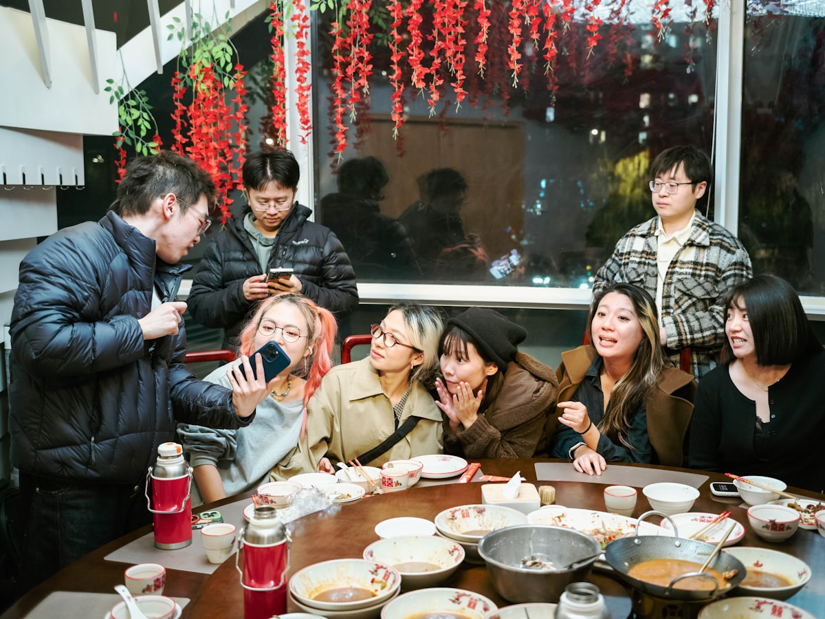 A group of friends gathered around a shared meal at a casual restaurant table, laughing together in warm evening light — the kind of after-work gathering where a hyung or noona title often gets earned