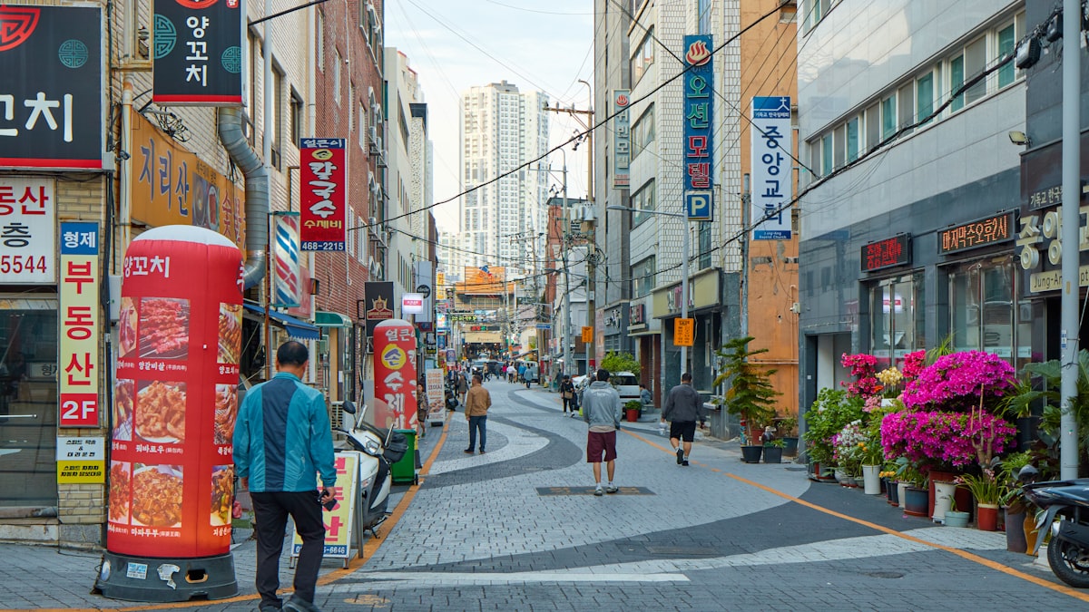 Korean commercial street lined with stacked shop signs, pedestrians, and potted azaleas
