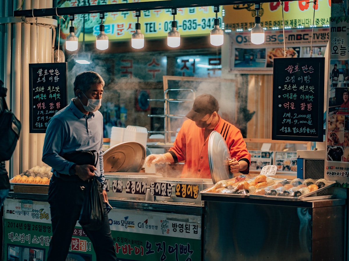 A steamy Korean street-food stall lit by hanging bulbs, a vendor cooking as a customer waits — the kind of traditional market setting where the May 2026 campaign controversy took place