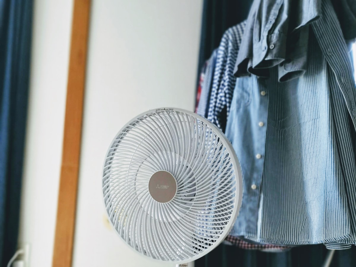 A small white electric fan running on the floor of a home interior, clothes hanging in the background