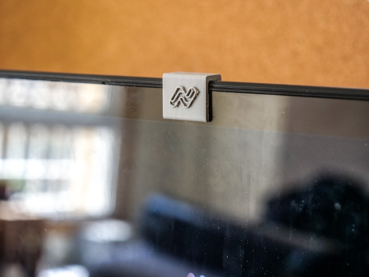 A small clip-on webcam cover attached over the camera at the top of a laptop or monitor screen, warm indoor light in the background