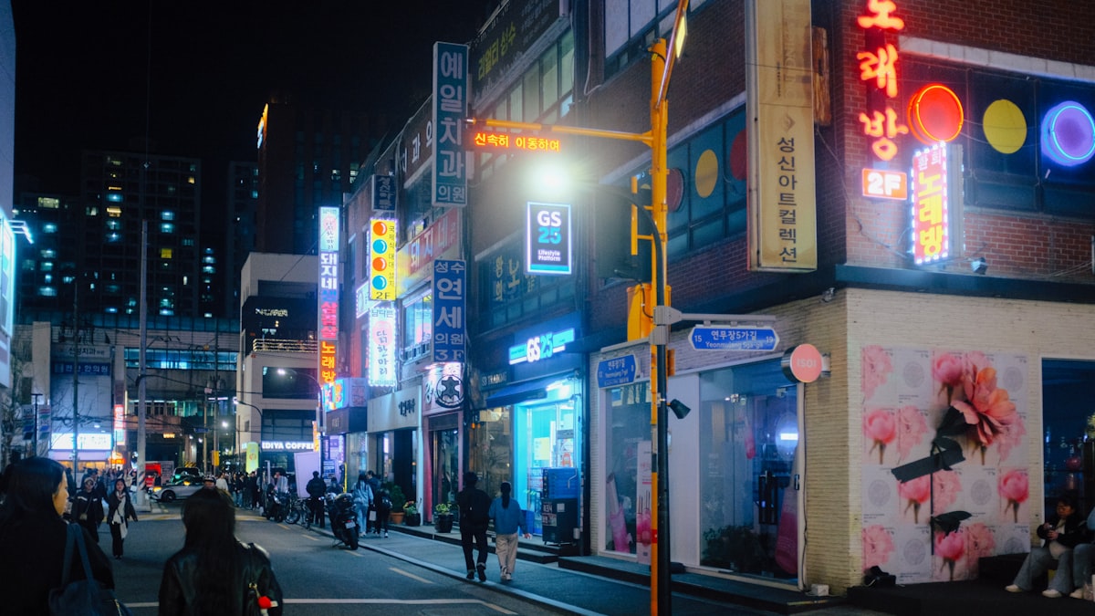 Korean street corner at night lit by neon signs for a noraebang, clinics, and a GS25