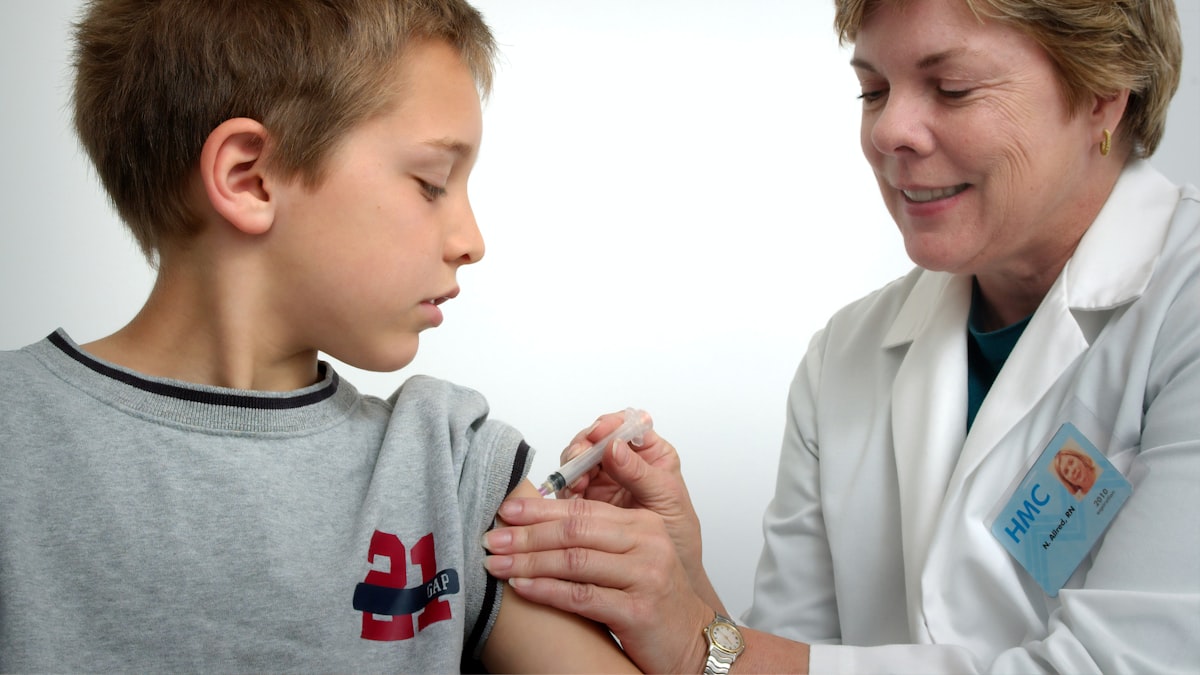 A nurse in a white coat administering a vaccine injection into a school-age boy's upper arm during a clinic visit