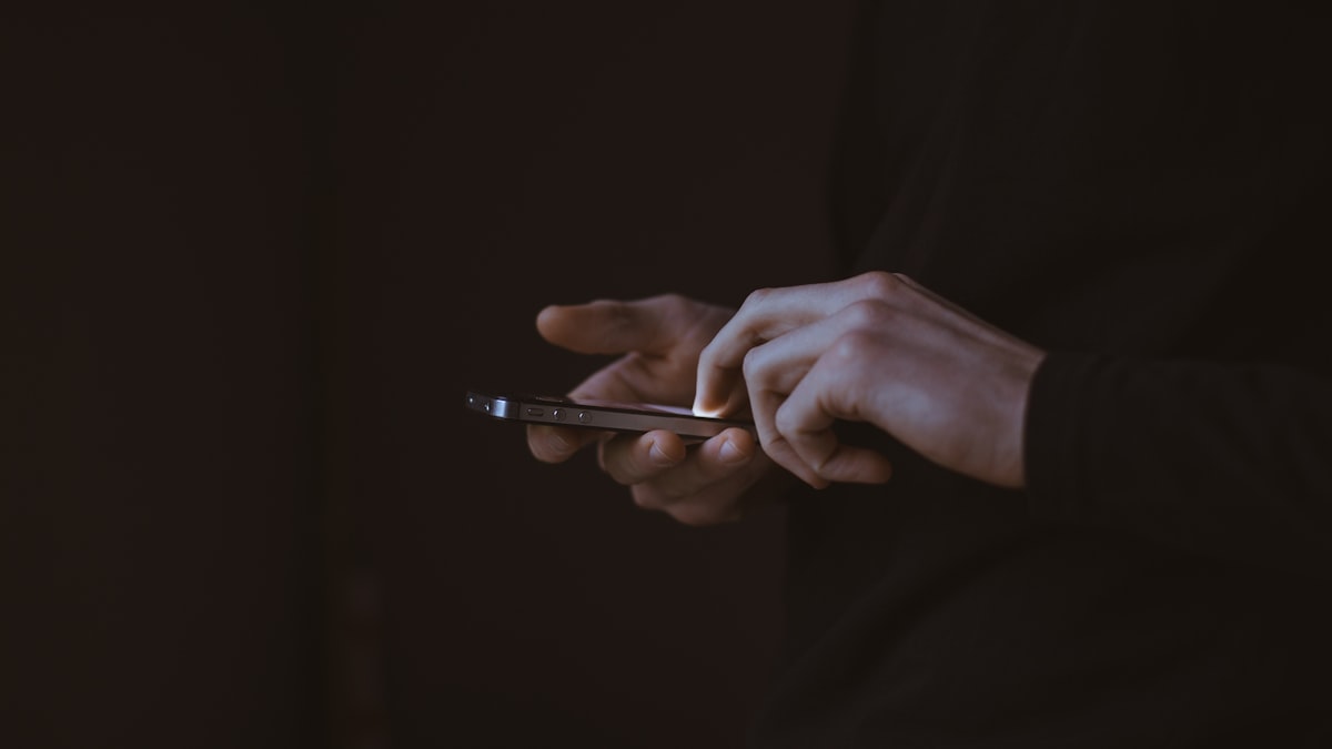 A person's hands checking a smartphone in near-total darkness, seen in side profile against a dark hoodie