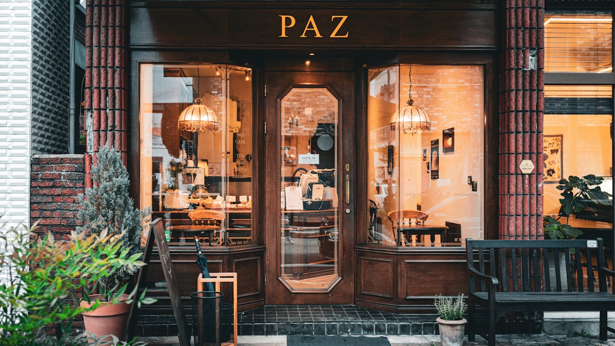 The warm-lit wooden entrance of a small cafe called PAZ at dusk, glass doors flanked by hanging pendant lights and potted plants