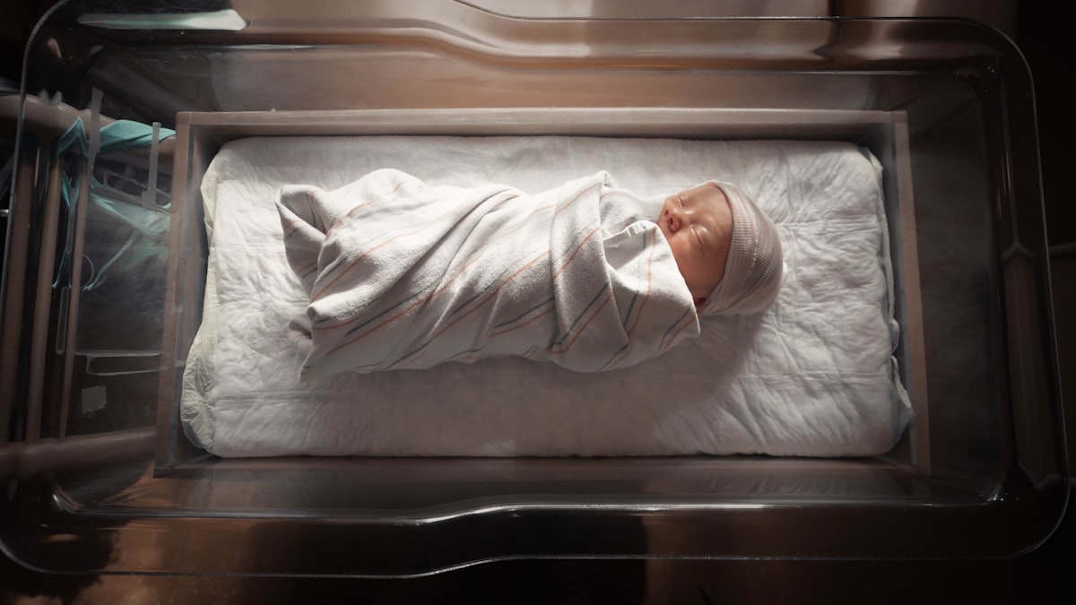 Newborn swaddled in a striped blanket, asleep in a clear hospital bassinet