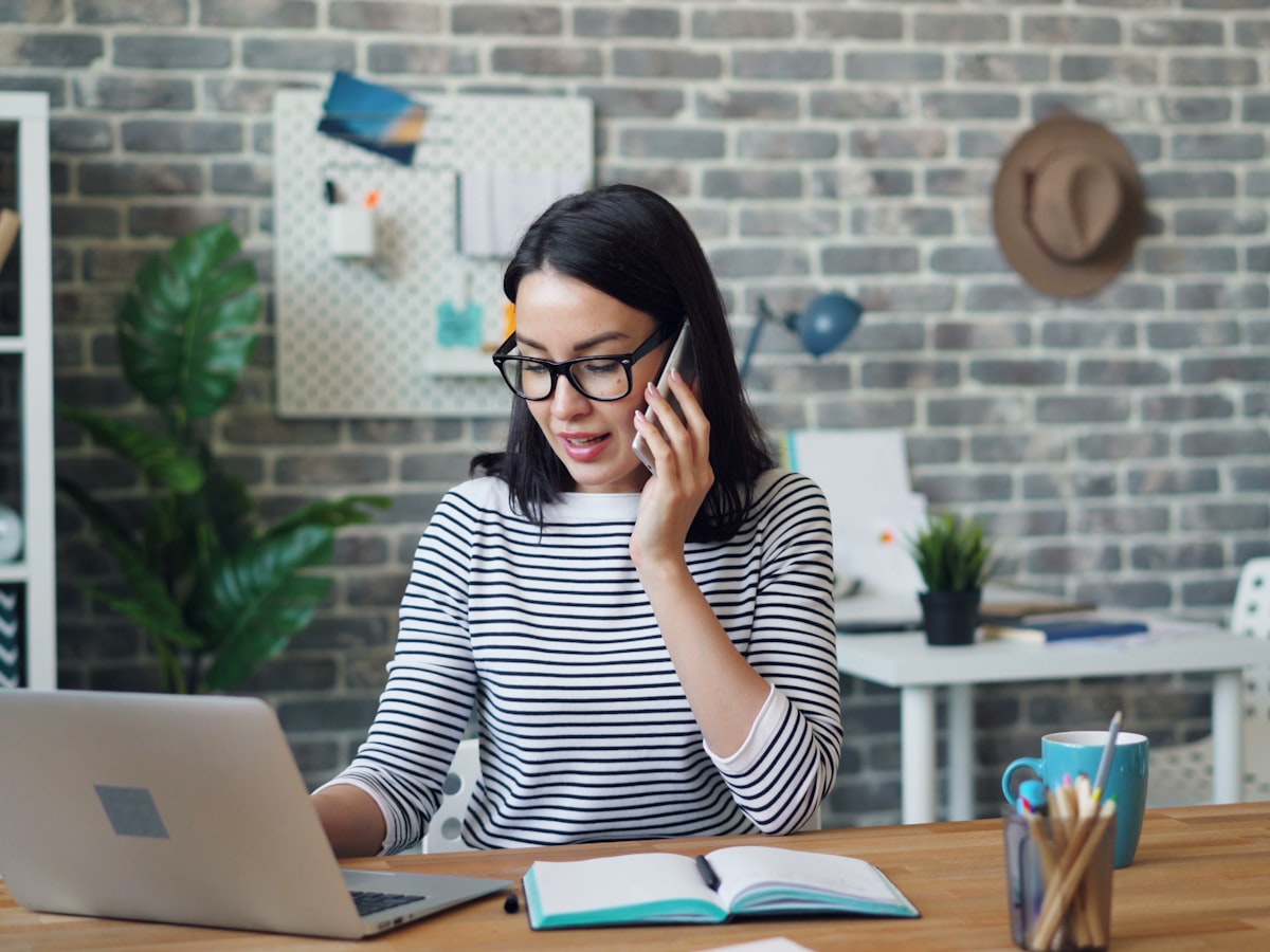 Woman on a phone call while looking at her laptop and notebook at a desk