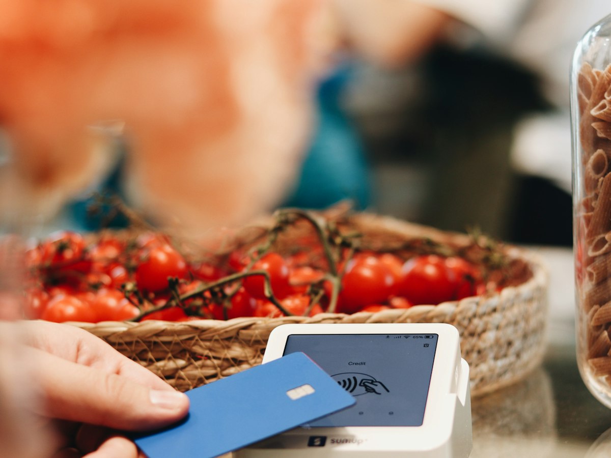 Close-up of a hand tapping a blue card on a payment terminal at a small food-stall counter, with a basket of tomatoes and a jar of pasta in the background