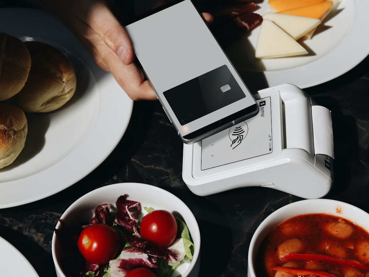 A hand holding a smartphone over a payment terminal at a restaurant table set with bread, cheese, salad, and soup