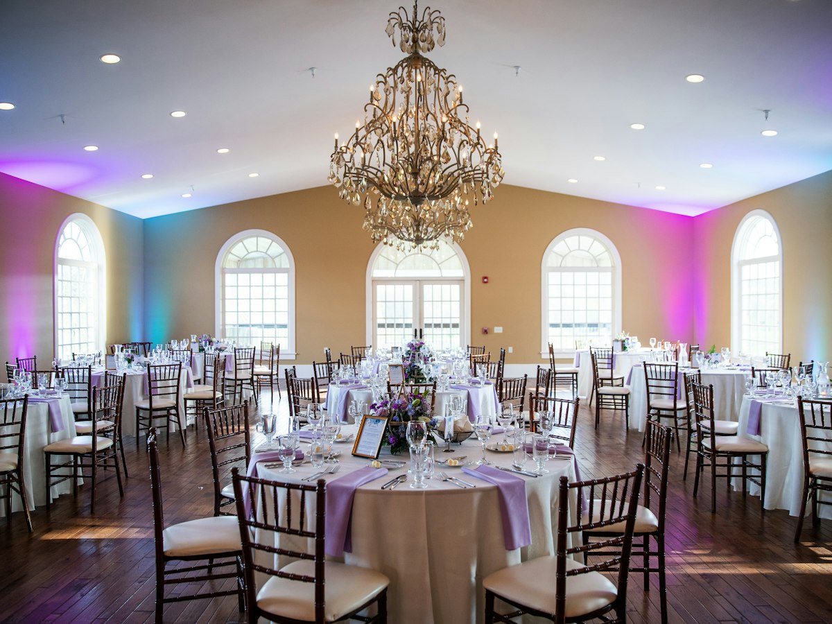 An elegant indoor banquet hall set up for a wedding reception, round tables with white linens and place settings under a crystal chandelier
