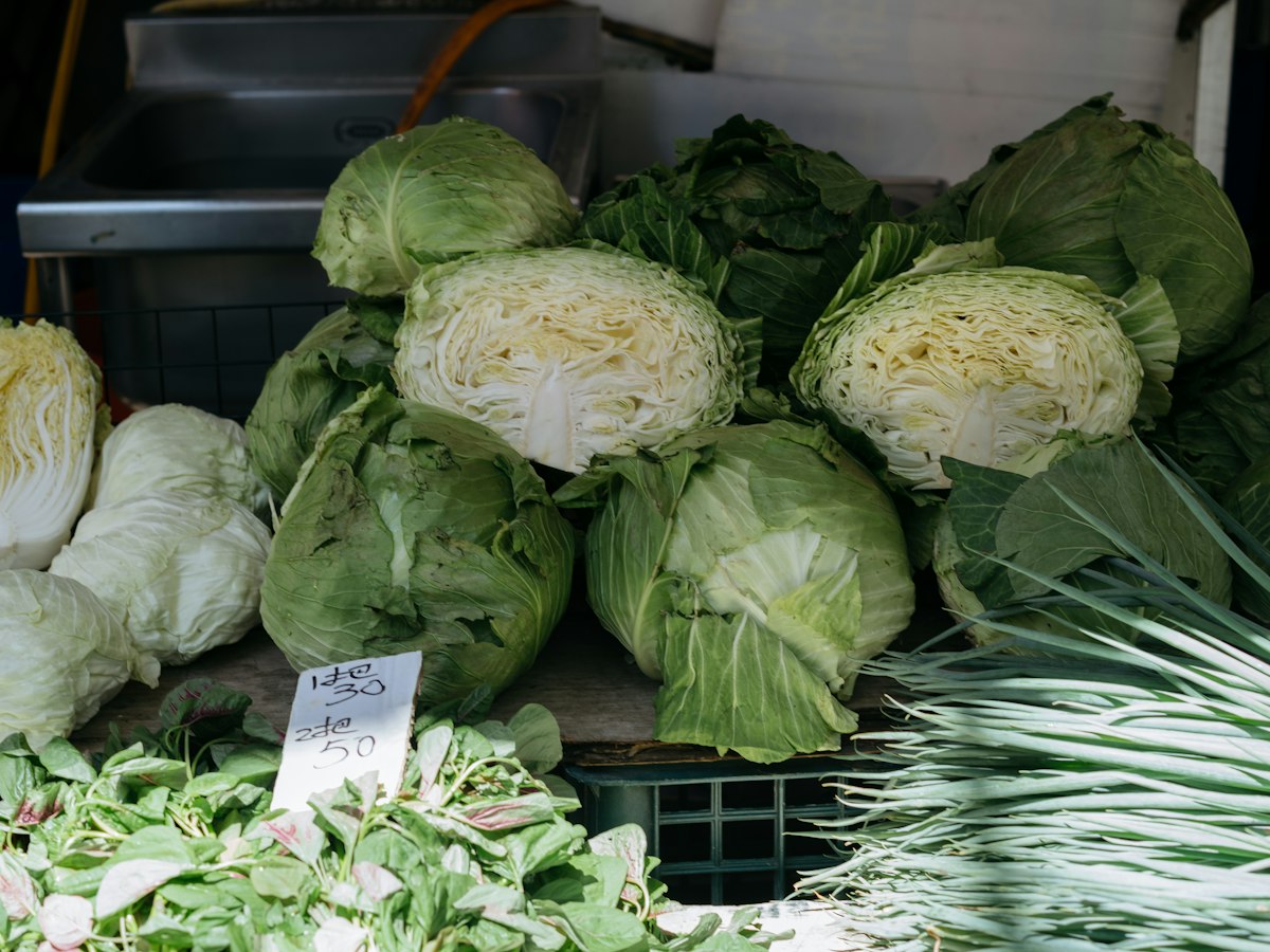 Cabbages and greens displayed at a produce market stall with a hand-written price sign