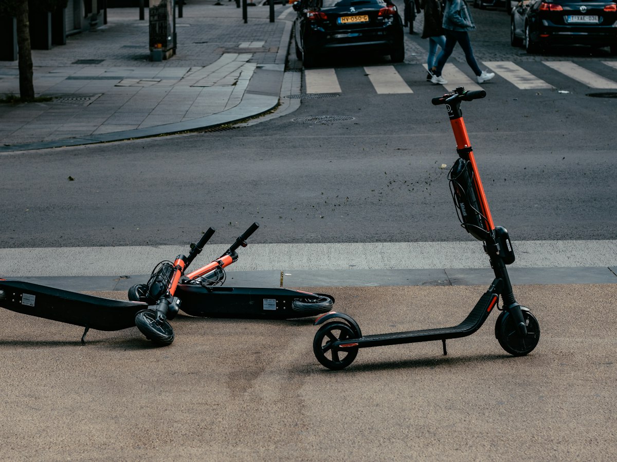 A shared electric kick scooter tipped over on a city sidewalk near a crosswalk as pedestrians pass by