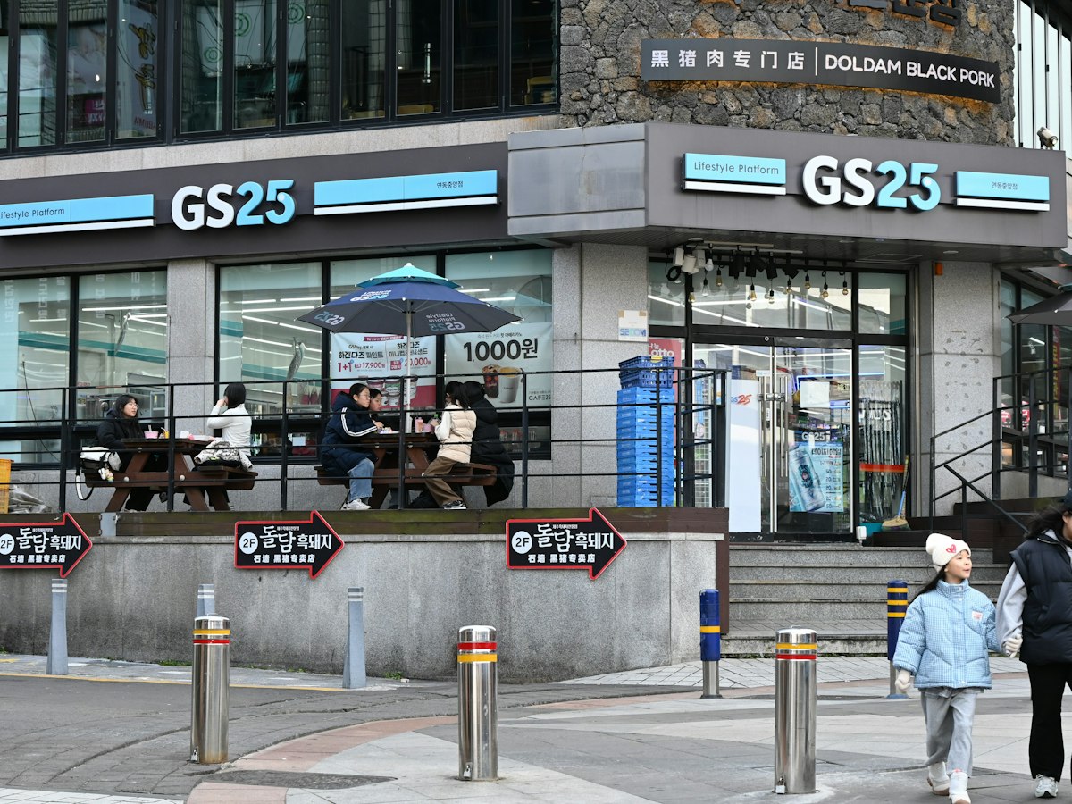 People eating at outdoor picnic tables outside a GS25 convenience store in Korea, with a 1,000-won price sign and store shelves visible behind them