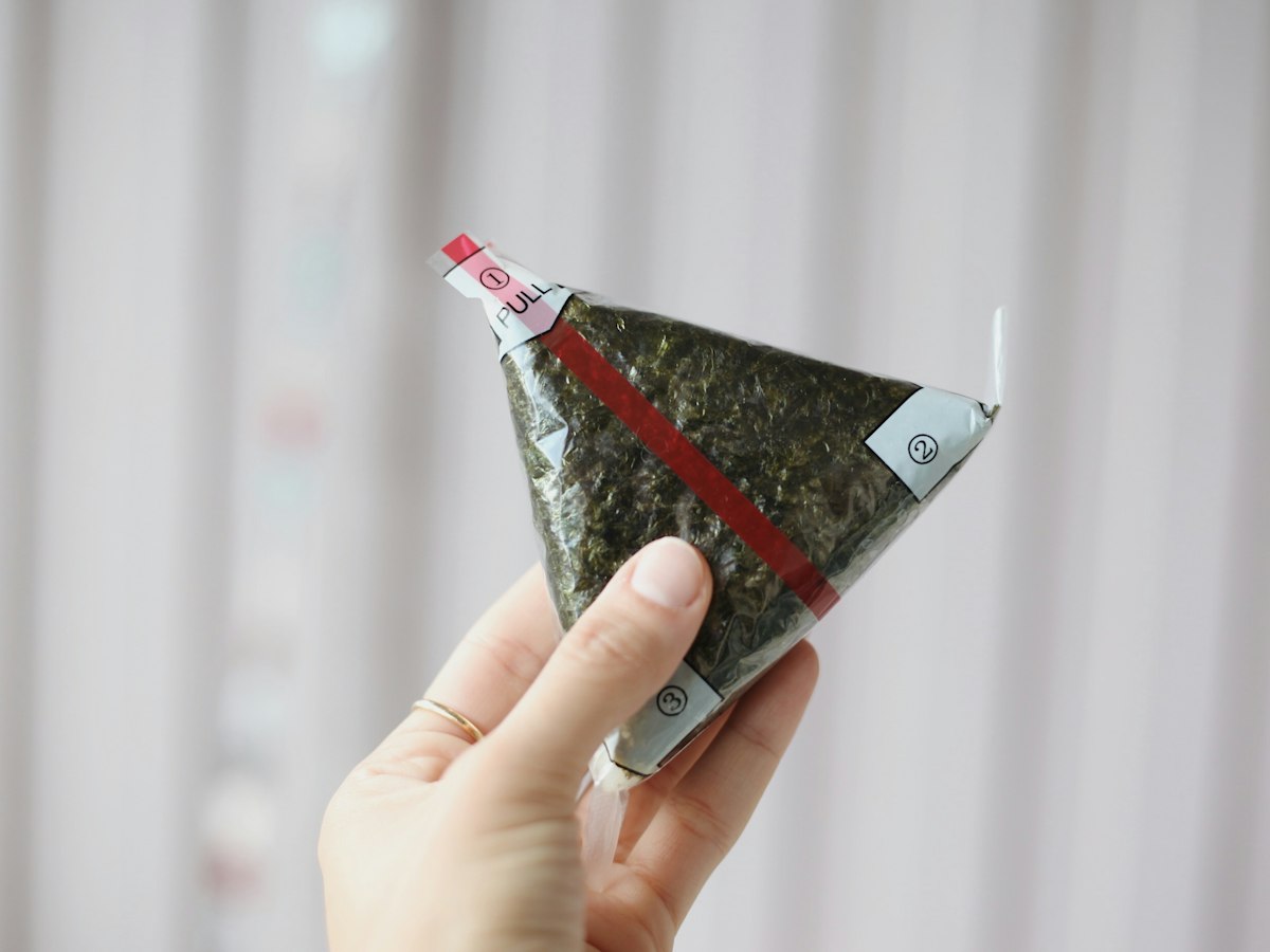 Close-up of a hand holding a sealed triangle-shaped rice ball (samgak gimbap / onigiri-style) snack, showing its numbered plastic pull-wrapper before opening