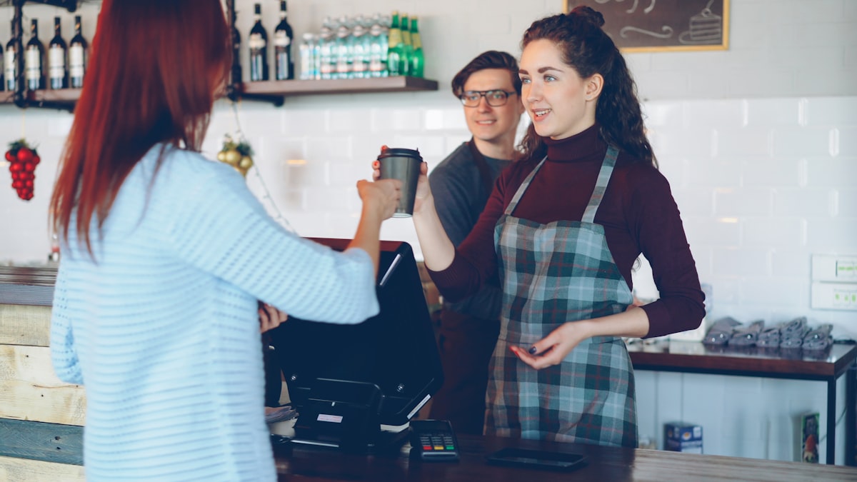A barista hands a takeout coffee cup across the counter to a customer, no cash or card changing hands