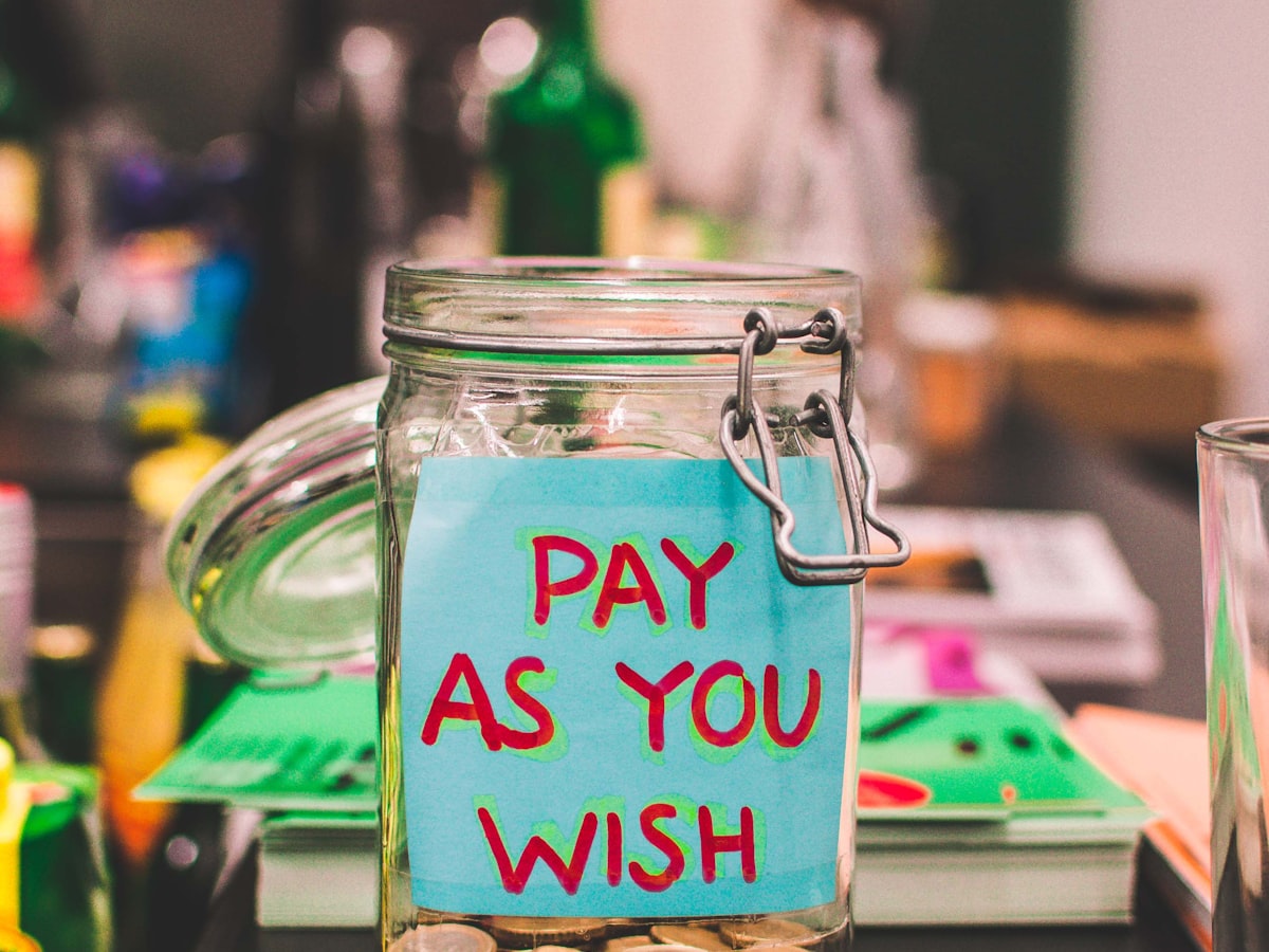 A glass jar with a handwritten Pay As You Wish sticker sitting on a counter, coins visible inside