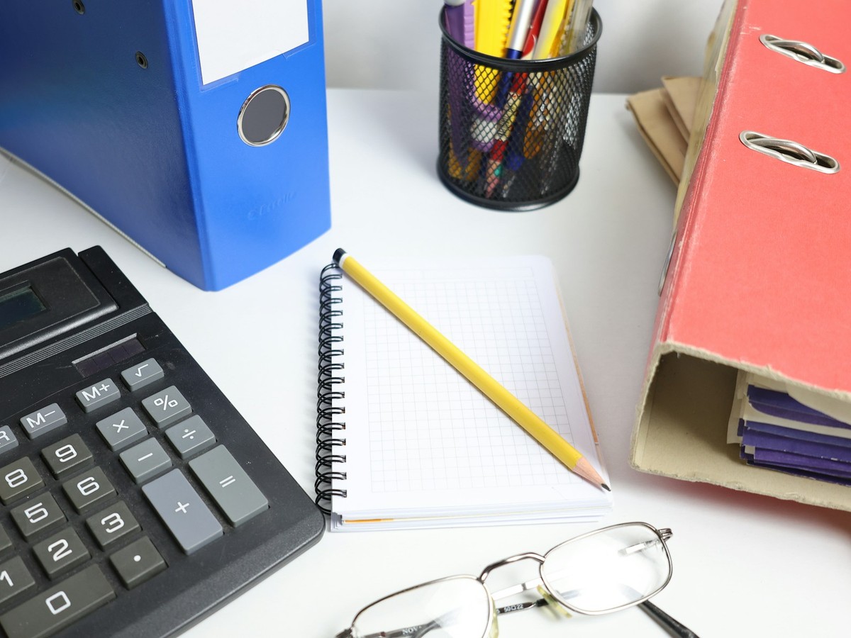 A calculator, a blank grid notebook with a pencil, office binders and reading glasses on a white desk