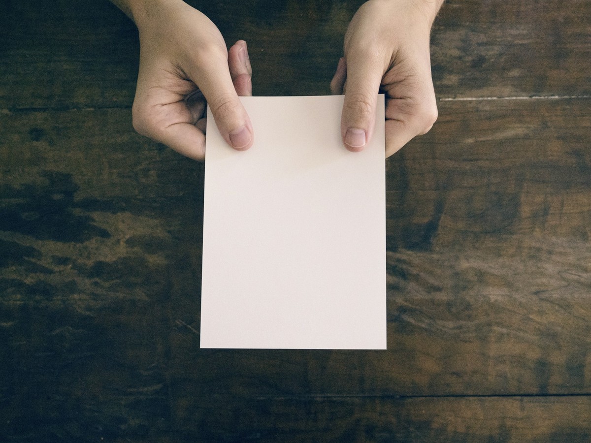Two hands holding a plain cream envelope over a dark wooden table, overhead view