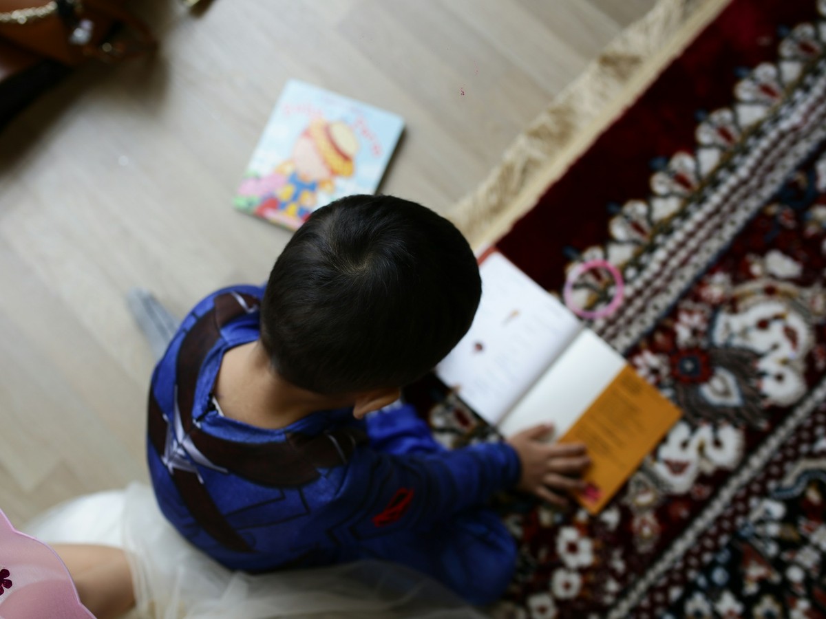 An Asian toddler with dark hair, seen from above, kneeling on a patterned rug and turning the pages of a picture book, a second picture book lying on the floor nearby